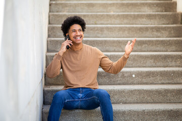 happy young hispanic man sitting on stairs talking with phone