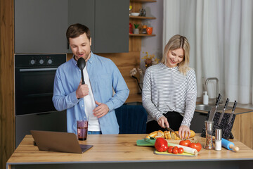 Home concert. Playful spouses having fun in kitchen, singing and dancing. Cheerful couple using kitchen utensils as microphones, fooling while cooking lunch at home.