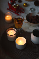 Cup of tea or coffee, glass of wine or juice, bowl of cookies, organic pomegranate, fluffy blanket, pine cones, various Christmas decorations and lit candles on the table. Christmas hygge concept.