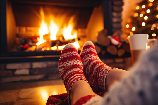 Feet In Woollen Socks By The Christmas Fireplace. Woman Relaxes By Warm Fire With A Cup Of Hot Drink And Warming Up Her Feet In Woollen Socks. Close Up On Feet. Winter And Christmas Holidays Concept.