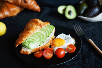 Croissant with avocado, scrambled eggs and cherry tomatoes on a black plate next to vegetables and a fork on a dark background