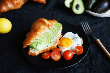 Croissant with avocado, scrambled eggs and cherry tomatoes on a black plate next to vegetables and a fork on a dark background