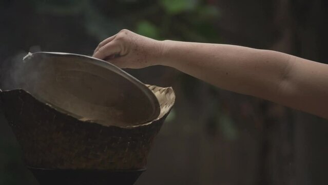 woman's hand opens the lid of a bamboo steamer that steams glutinous rice with smoke and hot steam.