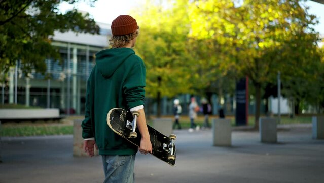 Trendy cool man holding skate board and looking away while walking at the urban park