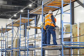 Workers men stand on construction scaffolds. Builders are installing insulation inside building. Process of interior finishing during construction. Men workers glue insulation material to walls.