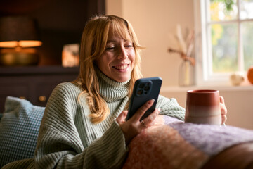 Woman At Home With Warming Hot Drink Of Tea Or Coffee In Cup Or Mug With Mobile Phone Streaming
