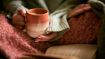 Close Up Of Woman At Home In Winter Jumper With Warming Hot Drink Of Coffee In Mug Reading Book