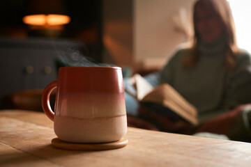 Close Up Of Hot Drink Of Tea Or Coffee In Cup Or Mug With Woman Reading Book In Background