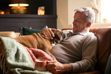 Mature Man At Home In Jumper With Hot Drink Of Tea Or Coffee In Cup Stroking Pet French Bulldog