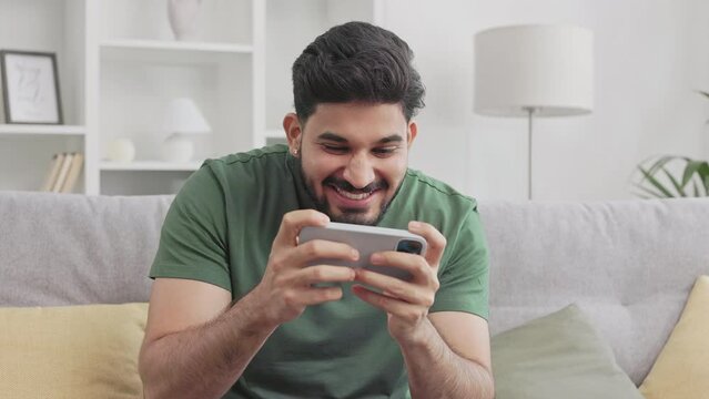 Bearded indian man winning in video games on modern smartphone while sitting on comfy couch at home. Young millennial guy in green t-shirt making yes gesture and sincerely rejoicing.