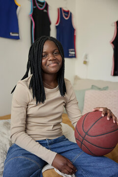 Smiling teenage girl with braided hair holding basketball while sitting in bedroom