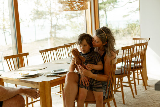 Mother with son holding teddy bear while sitting on chair by dining table