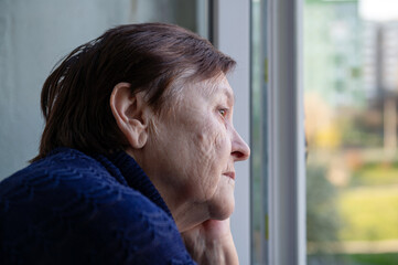 Portrait of a sad elderly woman looking out the window at home. A lonely elderly person looking out...