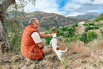 Mature gray haired man spending time outdoors with his small cute Jack Russell Terrier in mountain...