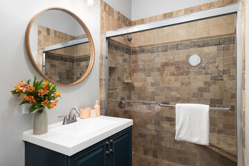 A bathroom with a blue cabinet wooden circular mirror above a white sink, and a shower with brown...