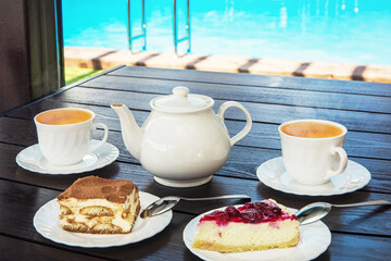 Afternoon tea set with hot tea and tasty cakes on the table and the swimming pool. Hotel resort or...