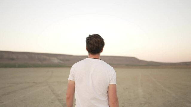 A young man in a white sweatshirt walks along the bottom of a dry lake against the backdrop of hills and begins to run. Back view