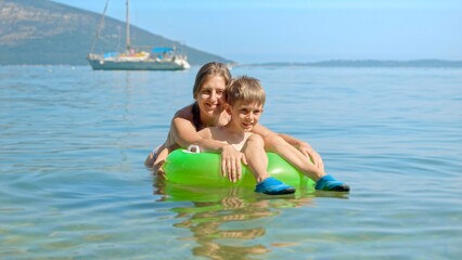 Cute boy sitting in inflatable swimming ring while mother is swimming next to him in the calm sea water at the beach. Family holiday, vacation and fun summertime of children and parents.
