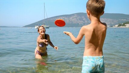 little boy throwing frisbee to his mother standing in the sea next to the beach. Family holiday, vacation and fun summertime of children and parents.