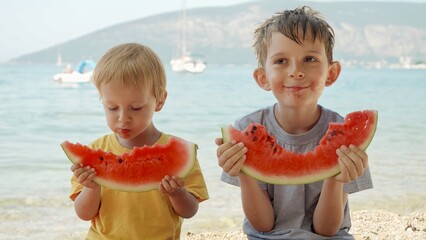 Two cute boys sharing sweet watermelon on a beach towel. Summertime fun, holiday happiness, and vacation relaxation