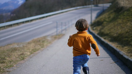From behind, a spirited child in a yellow pullover, jeans, and boots sprints with delight through...