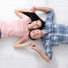 Fototapeta premium Top view of happy young couple looking at each other and smiling while lying on wooden floor. Woman and man holding hands and very happy