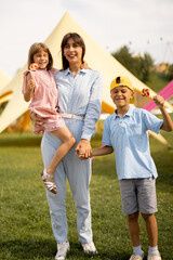 Fototapeta premium Happy mom with little girl and boy visit an amusement park, spending summertime happily together. Concept of children's entertainment and summer vacation