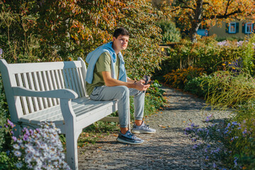 A Teenager Sits On A Bench In The Autumn Park Drinks Coffee From A Thermo Mug And Looks Into A Phone. Portrait Of Handsome Cheerful Guy Sitting On Bench Fresh Air Using Device Browsing Media Smm