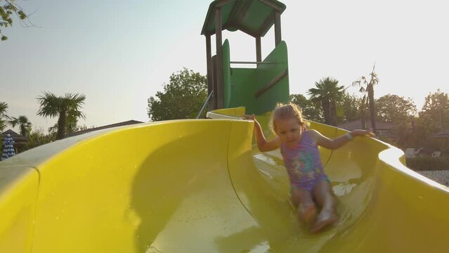 Cute, small girl sliding down a small yellow water slide at a swimming pool at the Bella Italia camp site in Peschiera del Garda in Italy. It is a hot and sunny summer day. Slow motion 4K video