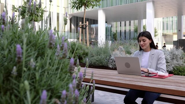 Young beautiful woman in smart casual clothes with laptop computer studying online sitting outside of his office building. Adult student listening training online business class on computer.