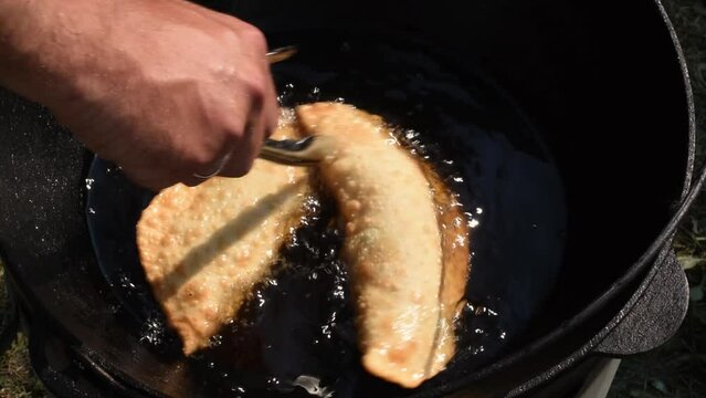A man fries pasties with meat in a cauldron, deep-fried in sunflower oil, he turns the cheburek with metal kitchen tongs. Dough in a frying pan with hot oil. Traditional food. Street food.