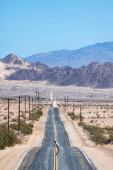 Young woman with hat and wearing shorts and a shirt walking in the middle of an asphalt road in Mojave Desert, California