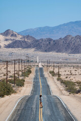 Young woman with hat and wearing shorts and a shirt walking in the middle of an asphalt road in Mojave Desert, California