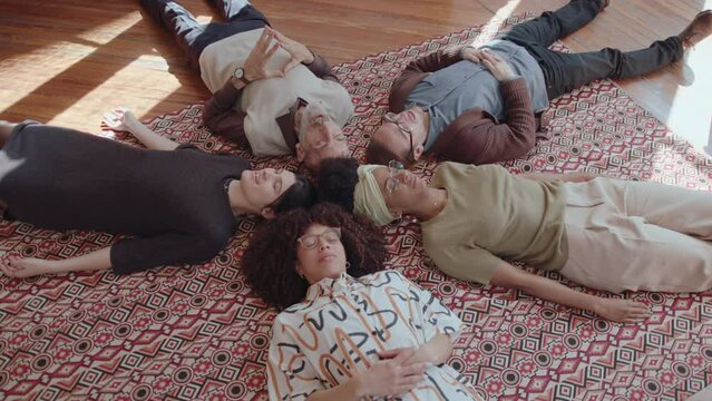 Group of therapy clients lying down on carpet with eyes closed, sharing their stories, having counseling session and supporting each other. High angle view