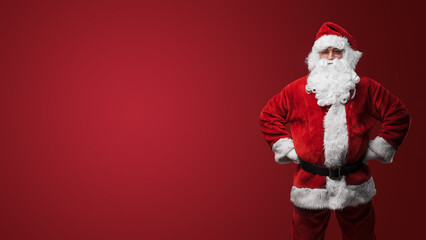 Jolly Santa Claus in traditional red suit standing with hands on hips against red backdrop