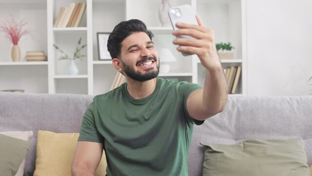 Smiling handsome man making thumb up, ok and peace signs while taking selfie on modern smartphone. Millennial guy sitting on grey couch and making new content for social media.