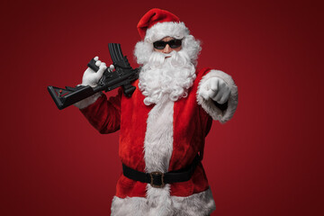 A man in a Santa Claus suit, wearing black sunglasses, poses with toy guns in hand against a red...