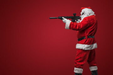 A man dressed as Santa Claus, wearing sleek black sunglasses, poses with toy machine guns against a bold red backdrop