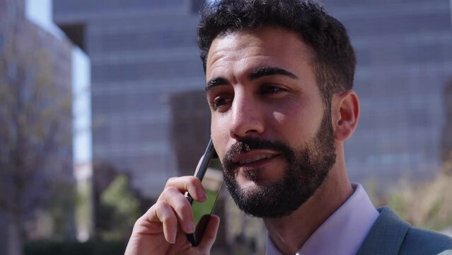 Young handsome Caucasian business man walking talking on phone serious expression. Male professional in suit receiving mobile call stroll outdoor. Successful worker with background of buildings.