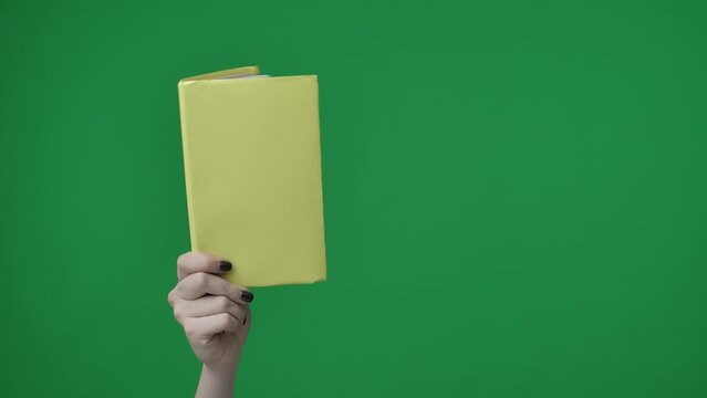 The frame features a close up of a woman on a green background. A woman's hand that holds an empty, rectangular, yellow A4 sheet of paper. She points it towards the camera.