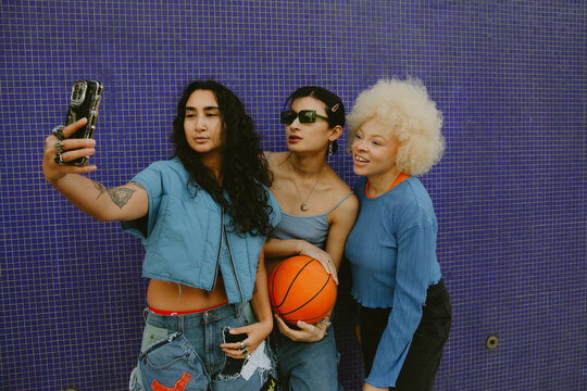 Three multiracial sporty friends in cool outfits taking selfie with basketball in front of blue wall outdoors