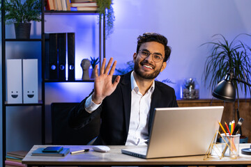 Hello. Indian business man working on laptop computer smiling friendly at camera and waving hands gesturing hi, greeting or goodbye, welcoming with hospitable expression at home office workplace desk