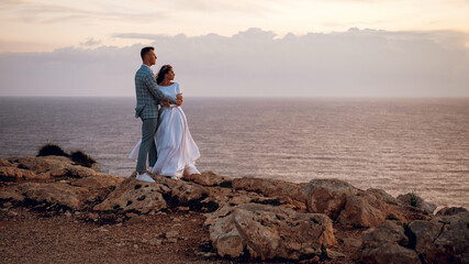 Gentle young wedding couple, bride and groom, hugging and enjoying the view on a rocky beach near the sea in evening Cyprus. Beautiful wedding photo shoot. Wedding for two, time for two.