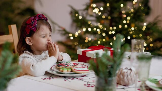 Christmas morning. Child eats sweets on New Year's Day. Toddler girl prays before eating and makes a wish. Cozy home with festive Christmas decorations and Christmas tree.
