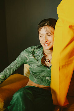 Young non-binary Asian person in fashionable green outfit posing on yellow couch in living room laughing