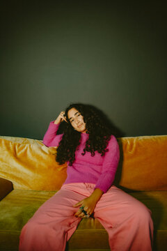 Young non-binary person wearing bright pink fashionable outfit sitting on yellow couch at home