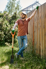 handsome farmer with beard wearing sun hat and standing with shovel near plants and fence