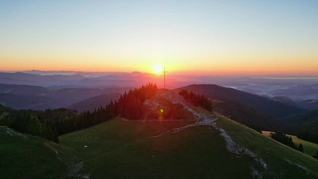 Drone flight into the sunrise passing a mountain summit cross in beautiful mountain range landscape in Styria, Austria