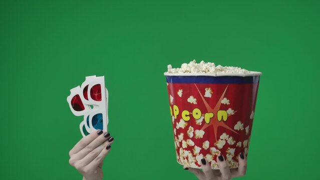 In the close up shot on the green background. A womans hand that is raised up and holds 3D glasses red blue and a box, a bucket of popcorn. Showing off to a movie theater for a movie