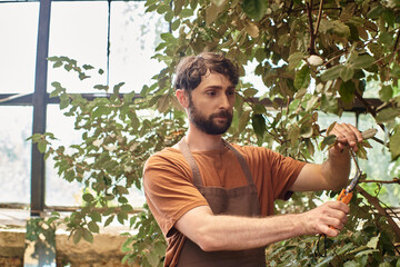 happy gardener in linen apron cutting branches of plants with gardening scissors in greenhouse